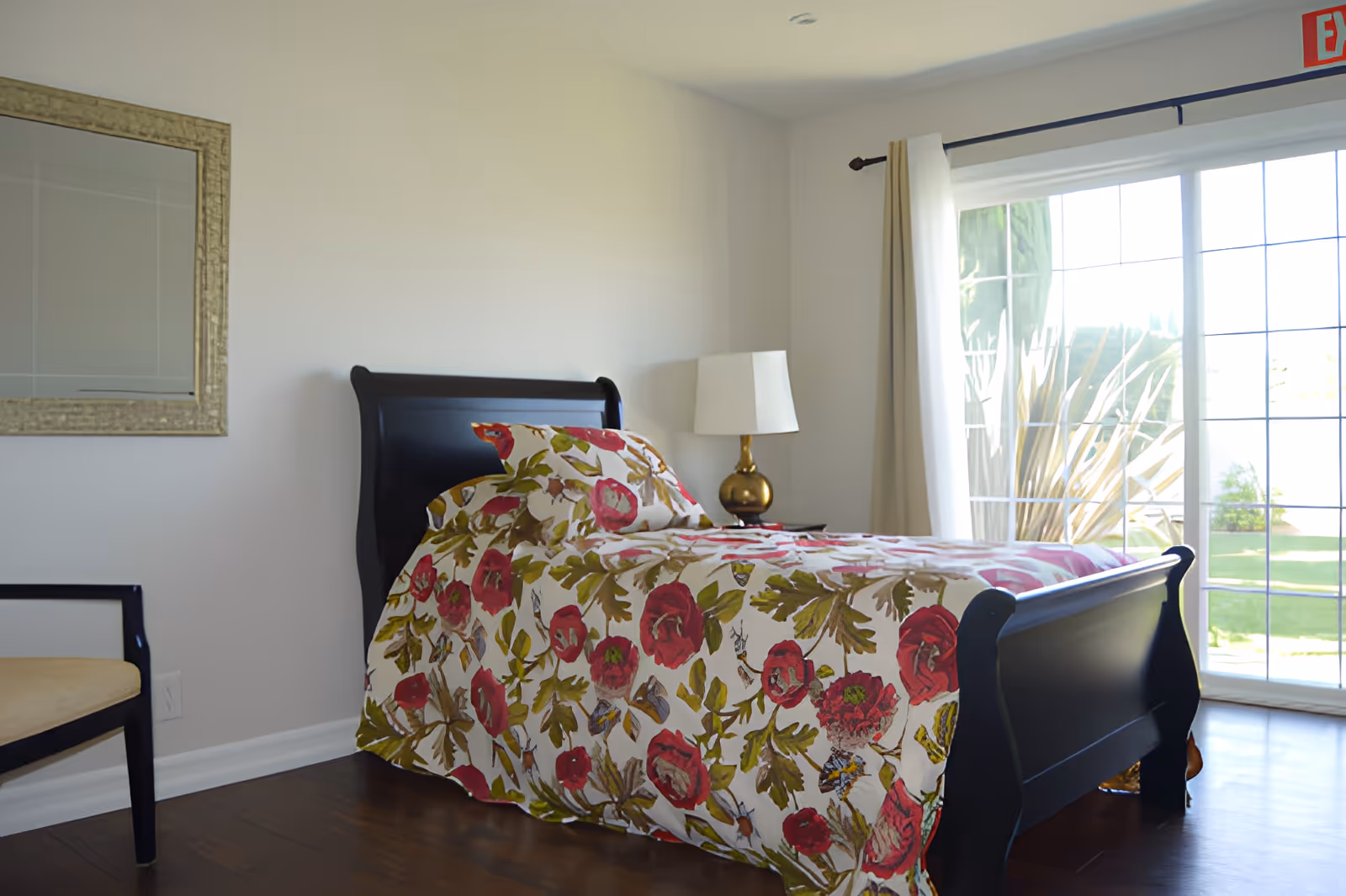 A bright bedroom with a dark wooden bed frame and a floral bedspread featuring red and green leaves. There is a gold-colored lamp on a nightstand next to the bed, a chair with a beige cushion, a large mirror on the wall, and a sliding glass door with curtains that opens to a green outdoor area.