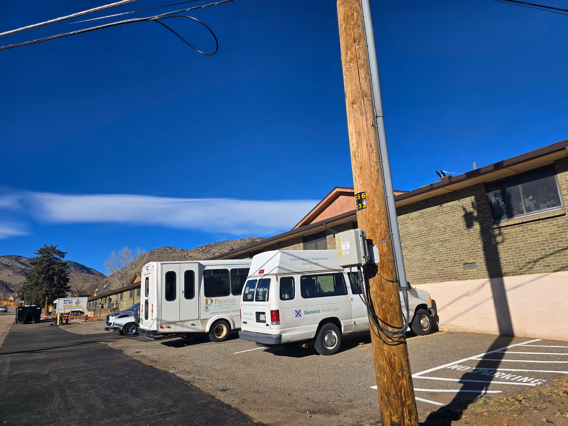 Parking lot outside a single-story brick senior care building with two white shuttle vans and a utility pole under a clear blue sky.