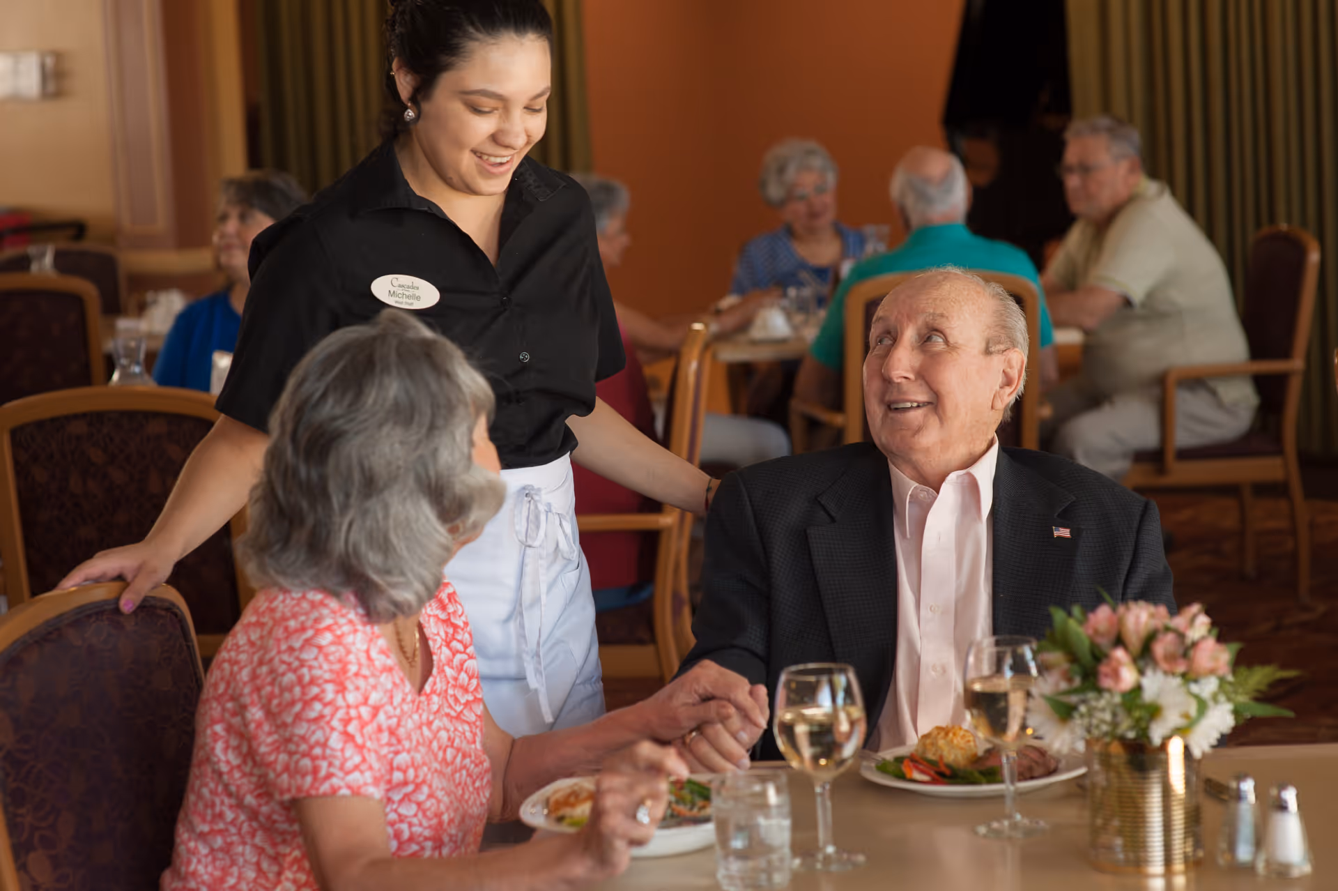 A smiling waitress named Michelle interacts warmly with an elderly couple seated at a dining table in a senior living facility. The elderly man is holding the woman's hand and looking up at the waitress. The table is set with plates of food, glasses of water and wine, and a small floral centerpiece. Other elderly residents are seated and dining in the background.