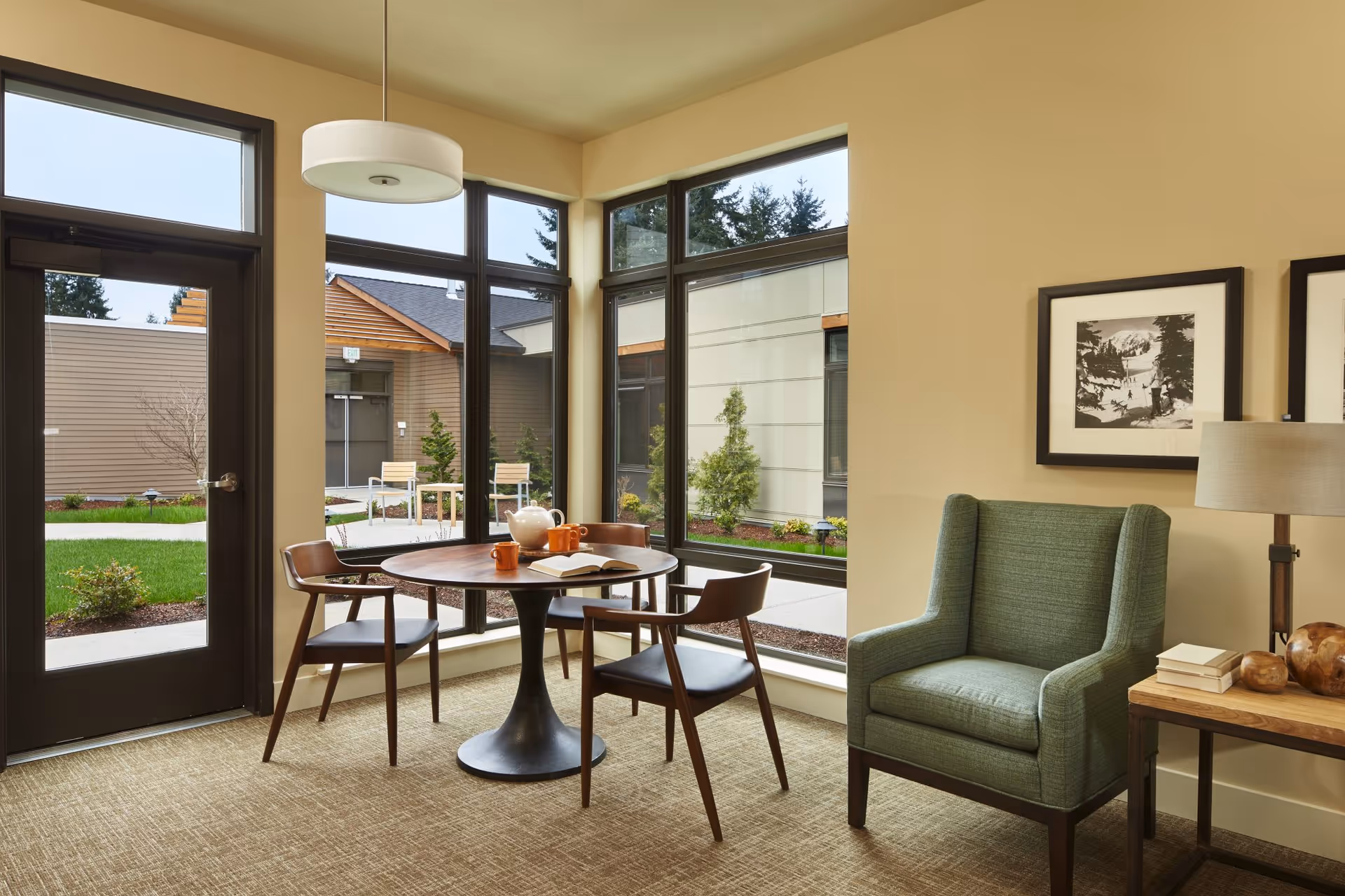 A cozy sitting area with a round wooden table surrounded by three wooden chairs with black seats. On the table, there is a teapot, two orange mugs, and an open book. Next to the table is a green upholstered armchair. A wooden side table with a lamp and some books is beside the armchair. Large windows and a glass door provide a view of a courtyard with greenery and outdoor seating. The walls are painted beige, and two black-and-white framed pictures hang on the wall above the armchair.