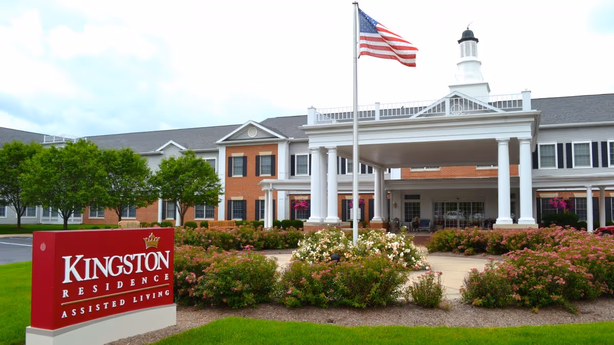 Front entrance of Kingston Residence assisted living with a red sign, flagpole, landscaped shrubs, and a covered porte-cochere.