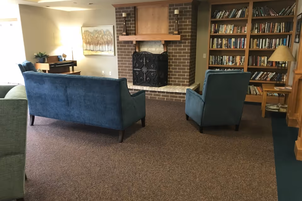 A cozy living room area featuring a blue sofa and matching armchair facing a brick fireplace with a decorative black screen. To the right is a wooden bookshelf filled with books and a table lamp. On the left side, there is a piano with a plant and a lamp beside it. The room has carpeted flooring and neutral-colored walls with a framed artwork above the piano.