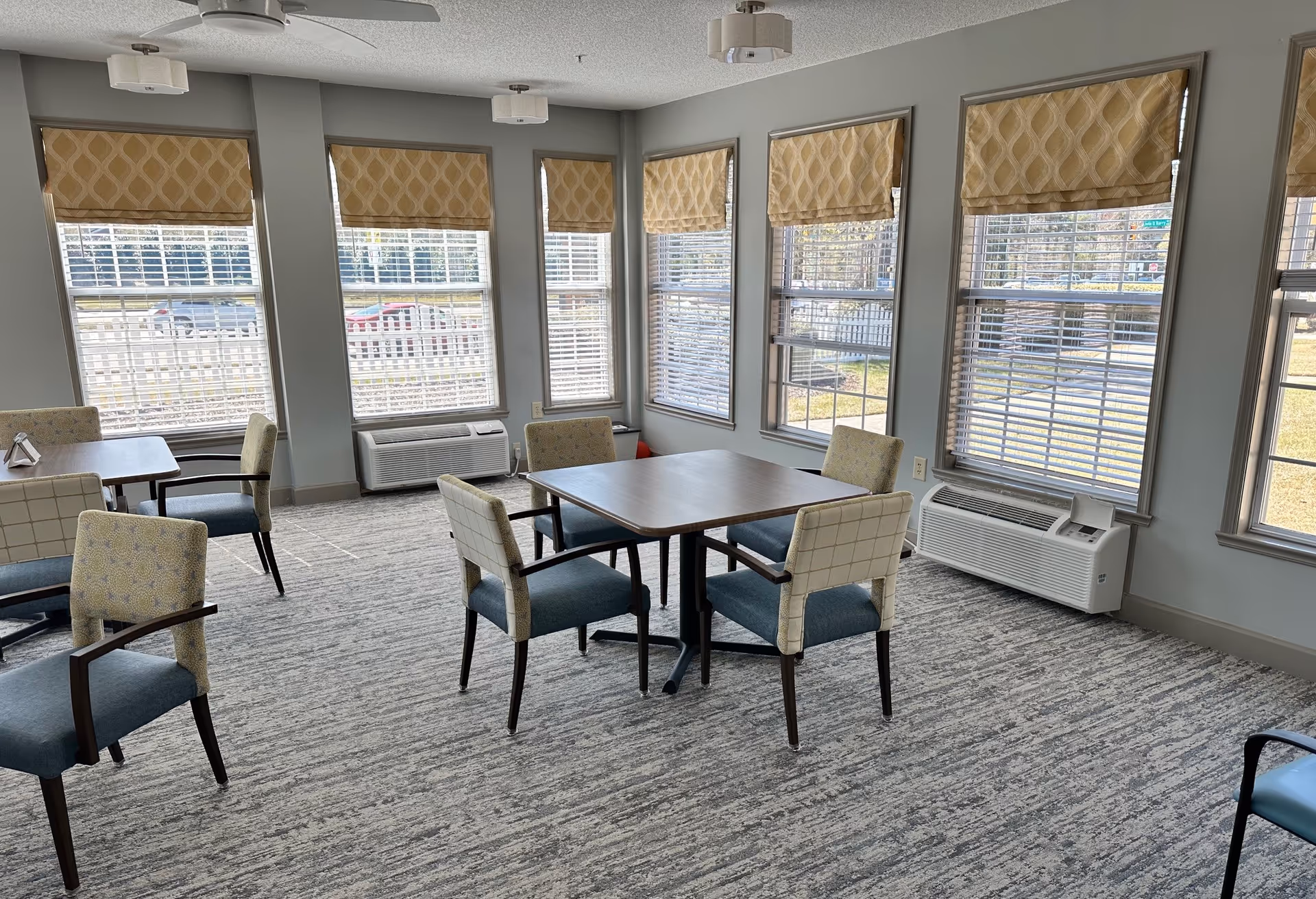A bright room with multiple windows covered by patterned beige Roman shades. The room has several square tables surrounded by cushioned chairs with blue seats and patterned backs. The floor is carpeted in a gray textured pattern, and there are two white air conditioning units beneath the windows. Ceiling lights and a ceiling fan are visible.