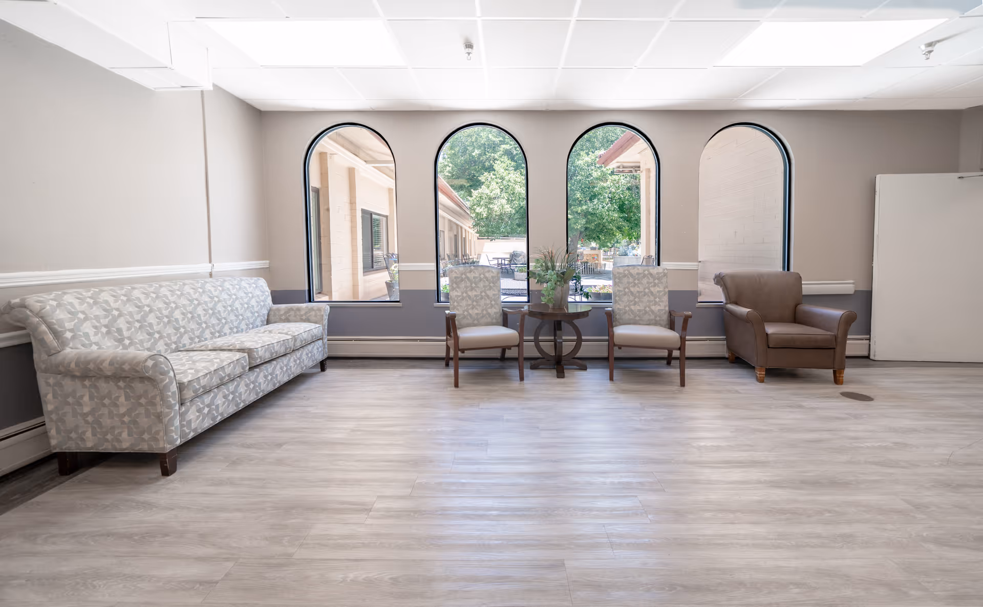 Bright seating area with a patterned sofa, three armchairs, a small round table with plants, and three arched windows looking outside.