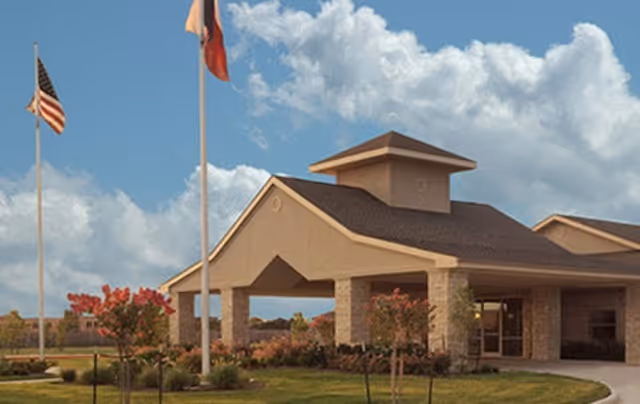 Entrance of a single-story senior living facility with a covered porte-cochère, flagpoles, and landscaped lawn under a partly cloudy sky.