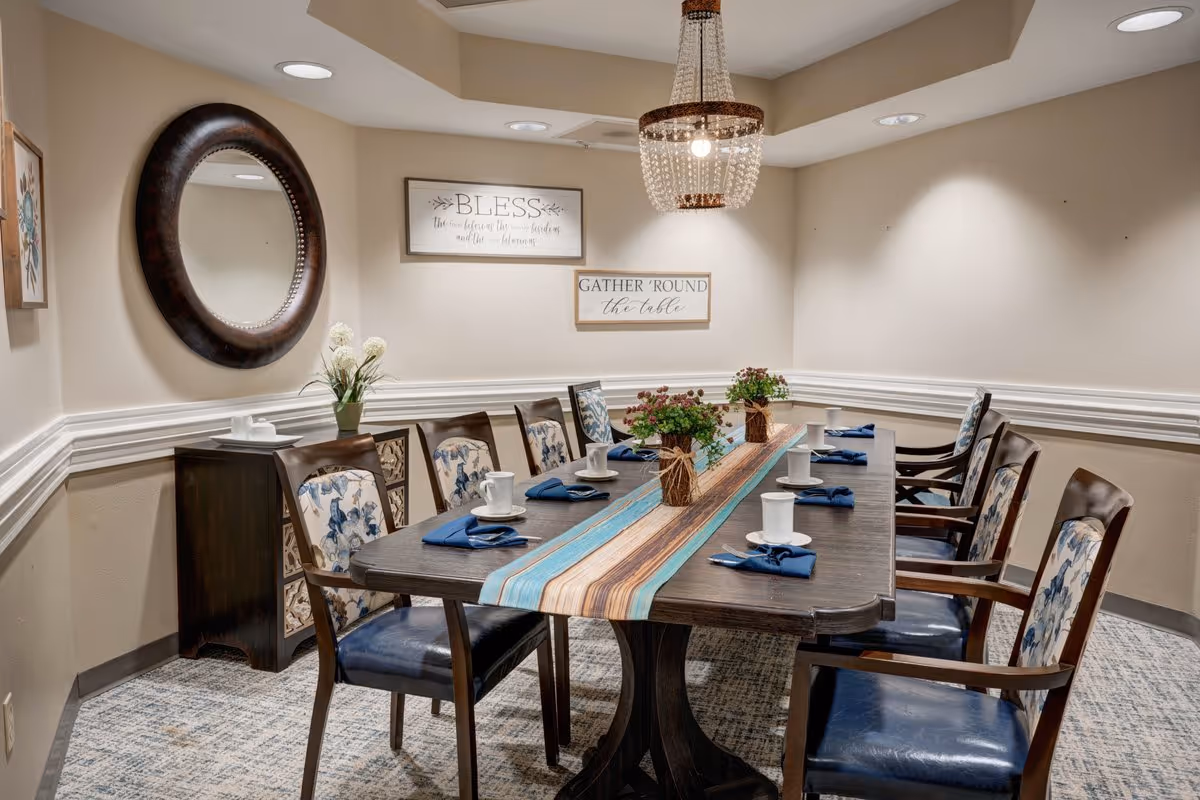 A dining room with a long rectangular wooden table set with white cups and blue napkins. The table has a decorative runner and two small flower arrangements. Eight wooden chairs with blue patterned cushions surround the table. The walls are beige with white trim, decorated with a round mirror and two framed signs that read 'BLESS the food before us, the family beside us, and the love between us' and 'GATHER 'ROUND the table'. A beaded chandelier hangs above the table.