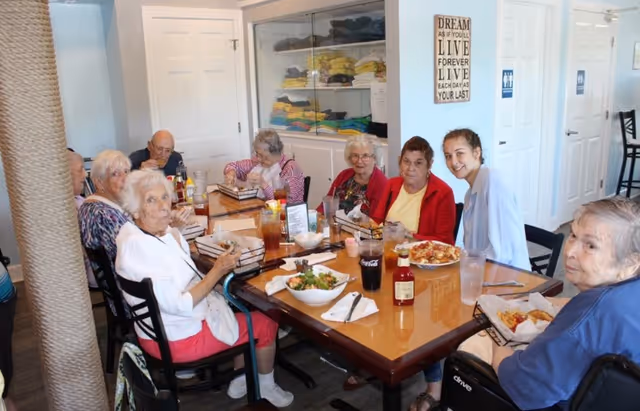 A group of elderly individuals and a caregiver sitting around a dining table enjoying a meal together in a bright room with white walls and a cabinet filled with folded towels in the background.