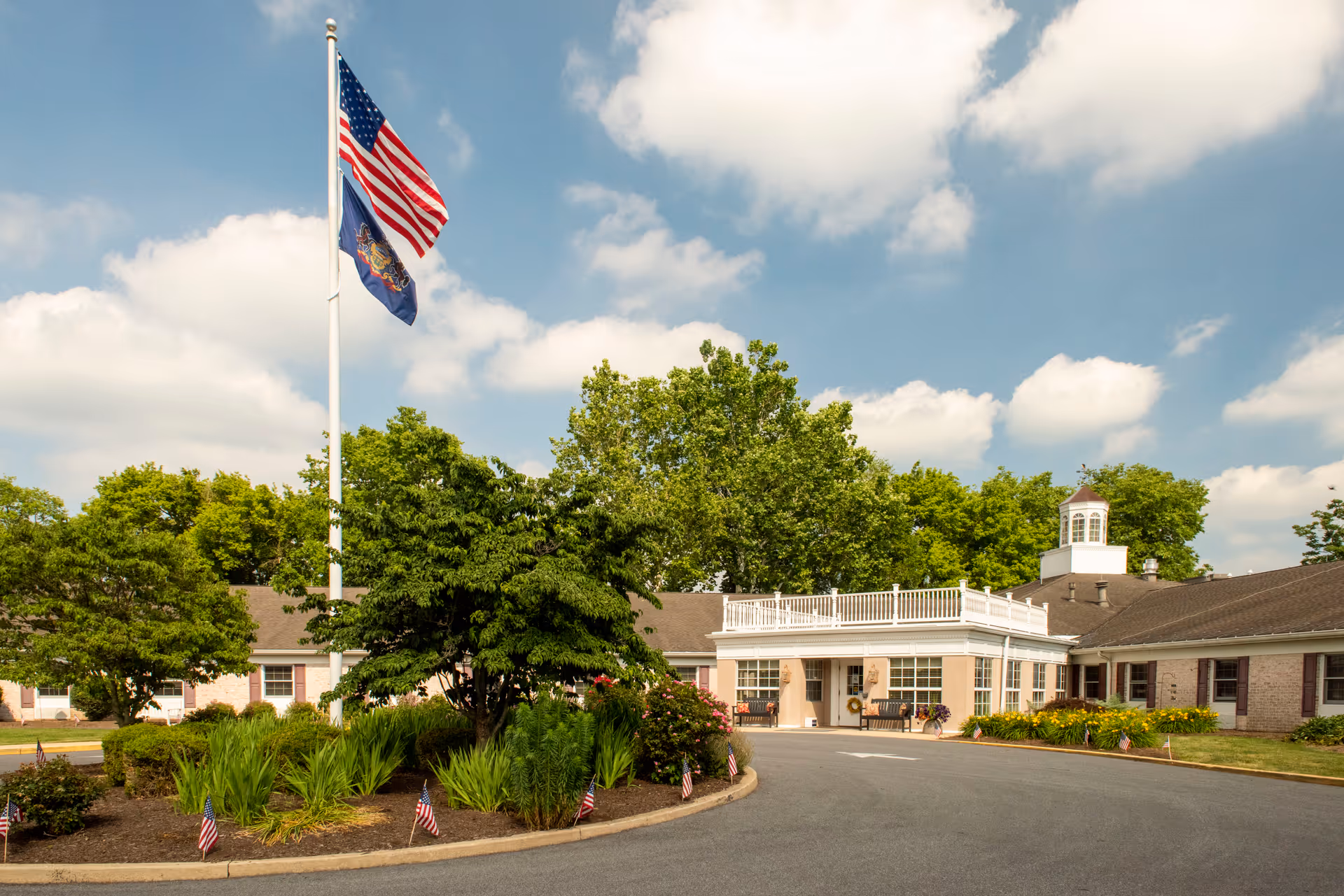 Exterior view of Providence Place Senior Living of Lancaster showing a single-story building with a circular driveway, landscaped greenery, and two flagpoles flying the American and Pennsylvania state flags under a partly cloudy sky.