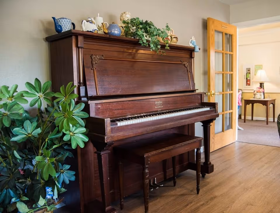 Upright wooden piano with a bench, potted plants, and decorative teapots in a bright senior living common room with a doorway to an adjacent sitting area.