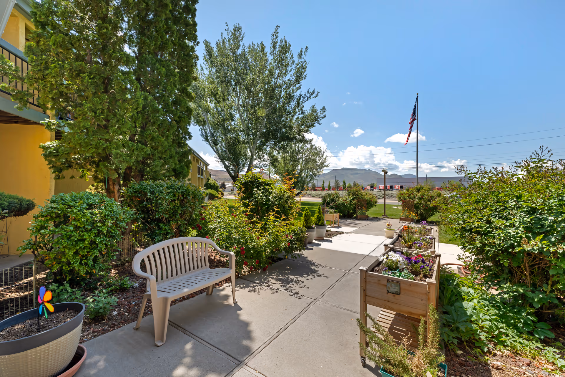 Outdoor garden area at Brookdale Sparks with a plastic bench, raised flower beds filled with colorful flowers, various green shrubs and trees, and an American flag on a flagpole against a blue sky with some clouds.