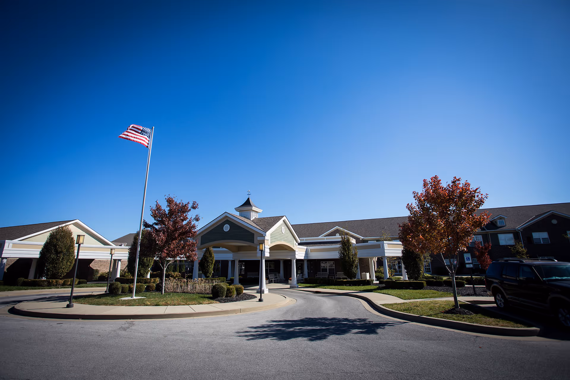 Exterior view of Bridgepointe at Ashgrove Woods, showing the front entrance with a covered driveway, landscaped greenery, trees with autumn foliage, an American flag on a flagpole, and a clear blue sky.