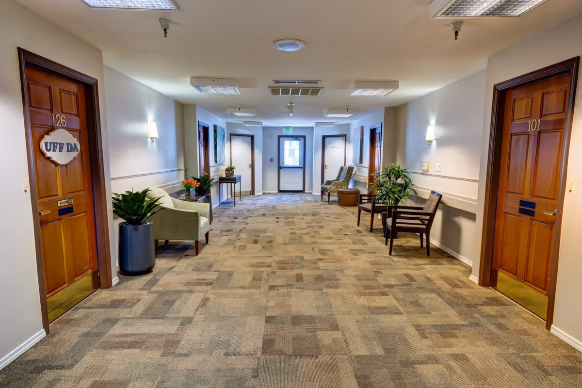 A well-lit hallway in a senior living facility with carpeted floor and beige walls. There are wooden doors on both sides labeled with room numbers 126 and 101. The door on the left has a sign that reads 'UFF DA'. The hallway is furnished with chairs, small tables, and potted plants. At the end of the hallway, there is a door with a window letting in natural light.