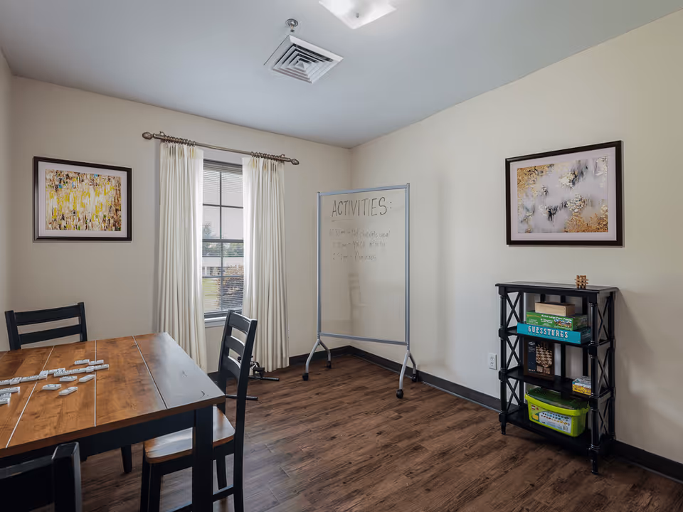 A well-lit room with a wooden table and black chairs, a window with cream curtains, a whiteboard listing activities, and a black shelving unit holding board games and puzzles. Two framed abstract paintings hang on the walls.