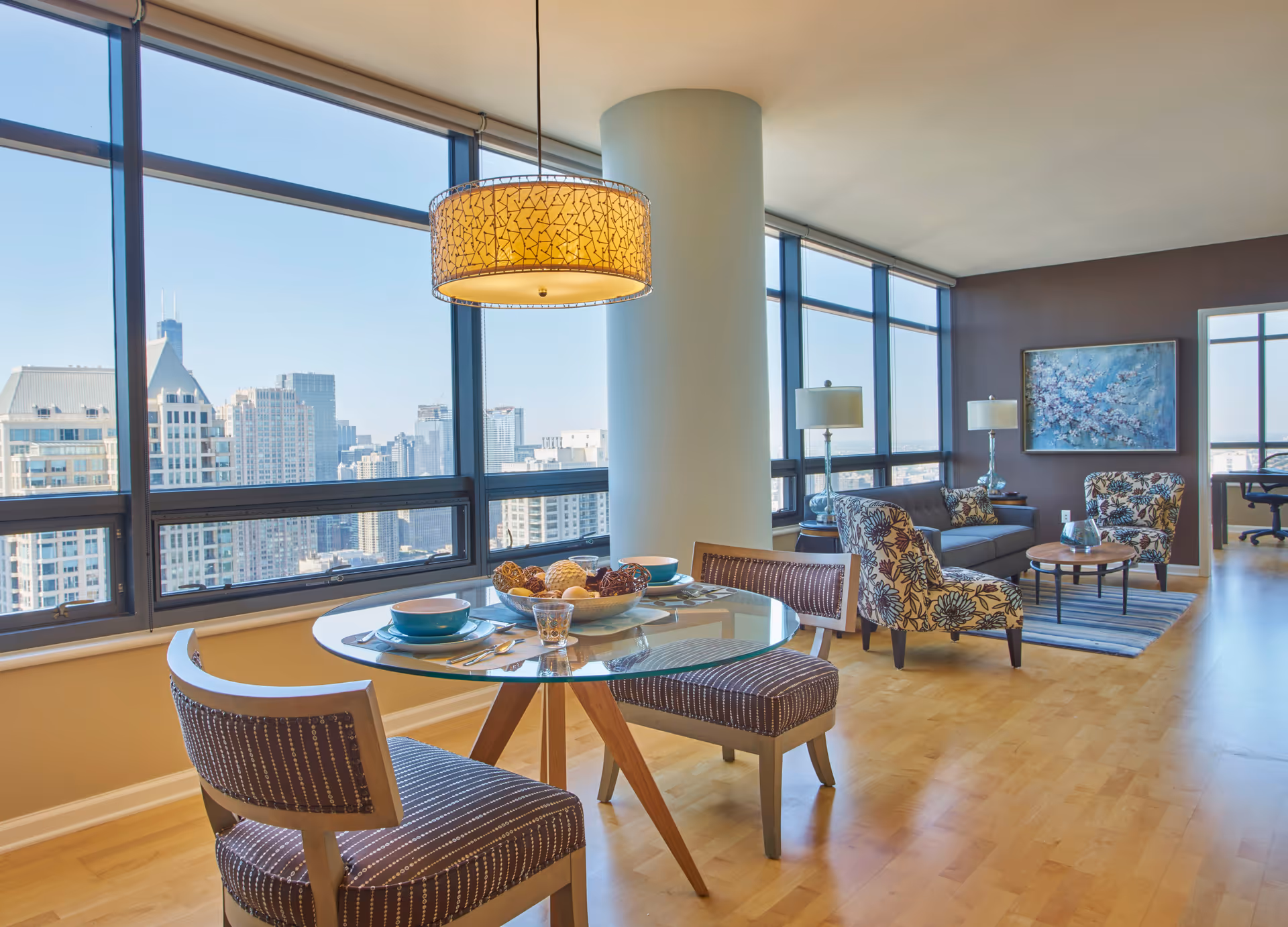 A bright and modern living and dining area in a high-rise apartment with large windows offering a city skyline view. The dining area features a round glass table set with bowls, plates, and utensils, surrounded by two upholstered chairs. The living area includes a gray sofa, two floral-patterned armchairs, a round coffee table, two table lamps, and a large painting on the wall. Light wood flooring and a large cylindrical pillar are also visible.
