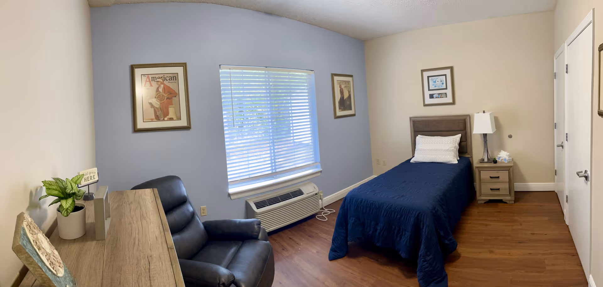 A small, neatly arranged bedroom with a single bed covered in a dark blue quilt and a white pillow. Next to the bed is a wooden nightstand with a lamp and a box of tissues. There is a black leather recliner chair and a wooden desk with a small plant and decorative items. The room has light-colored walls with one wall painted blue, three framed pictures, a window with blinds, and a wooden floor.