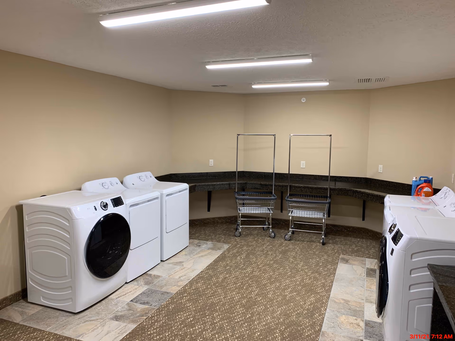 A laundry room with four white washing machines and dryers, two metal laundry carts with wheels, beige walls, a speckled brown carpet, and a long dark countertop running along the back and right walls. Laundry detergent is placed on the countertop.