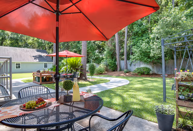 Patio with a round metal table set with fruit and a pitcher beneath a red umbrella, overlooking a grassy yard with a curved walkway and trees.