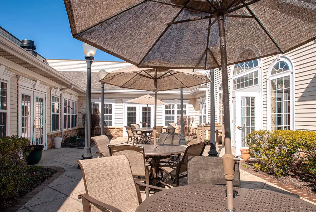 Sunlit outdoor courtyard with round patio tables, umbrellas and chairs in front of a beige memory care building.