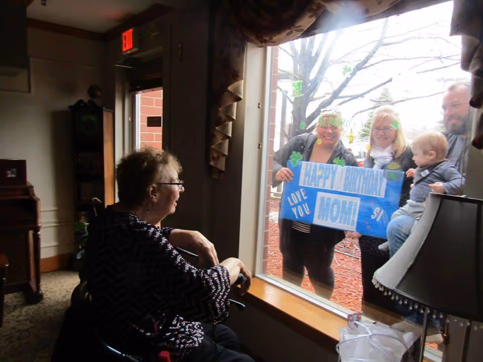 An elderly woman sitting inside near a window, looking outside at three adults and a child who are holding a blue sign that says 'Happy Birthday Mom! Love You'. The people outside are smiling and appear to be celebrating the woman's birthday from outside the window.