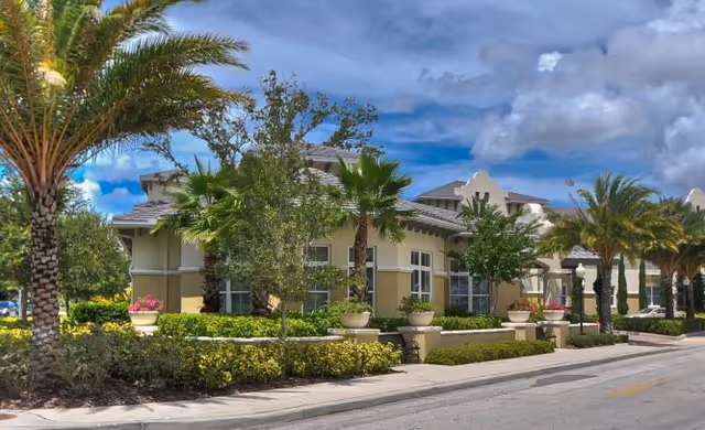 Exterior view of The Estate at Hyde Park facility showing a single-story building with beige walls and large windows, surrounded by palm trees, shrubs, and other greenery under a partly cloudy blue sky.