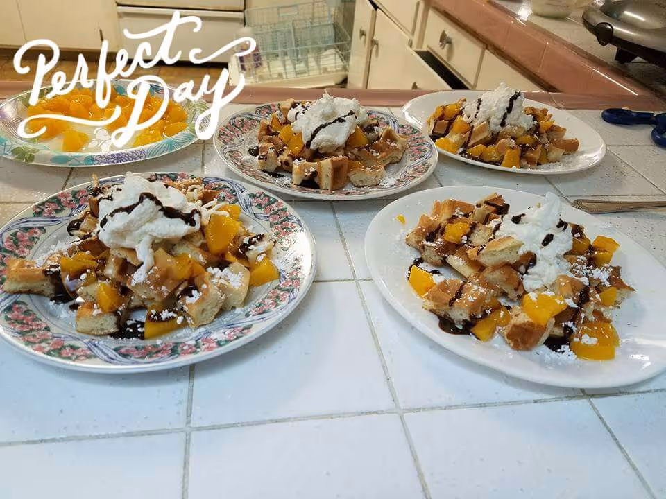 Plates of waffles topped with whipped cream, peach slices, chocolate drizzle and powdered sugar on a tiled kitchen countertop.
