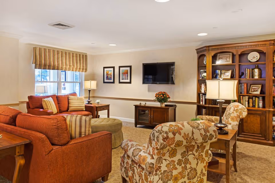 A cozy living room with orange sofas, a floral armchair, a round ottoman, wooden side tables with lamps, a wall-mounted flat-screen TV, framed artwork, and a large wooden bookshelf filled with books and decorative items.