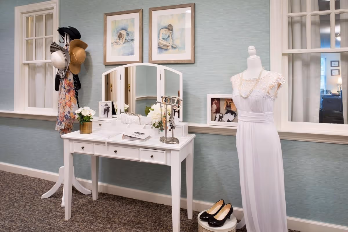 A white vanity table with a tri-fold mirror, hats on a coat rack, floral accents, and a mannequin dressed in a white gown against a pale blue wall.