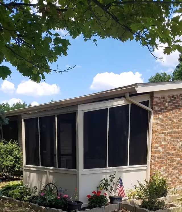 A screened-in porch attached to a brick building, surrounded by a small garden with various plants and flowers, under a blue sky with some clouds and tree branches overhead.