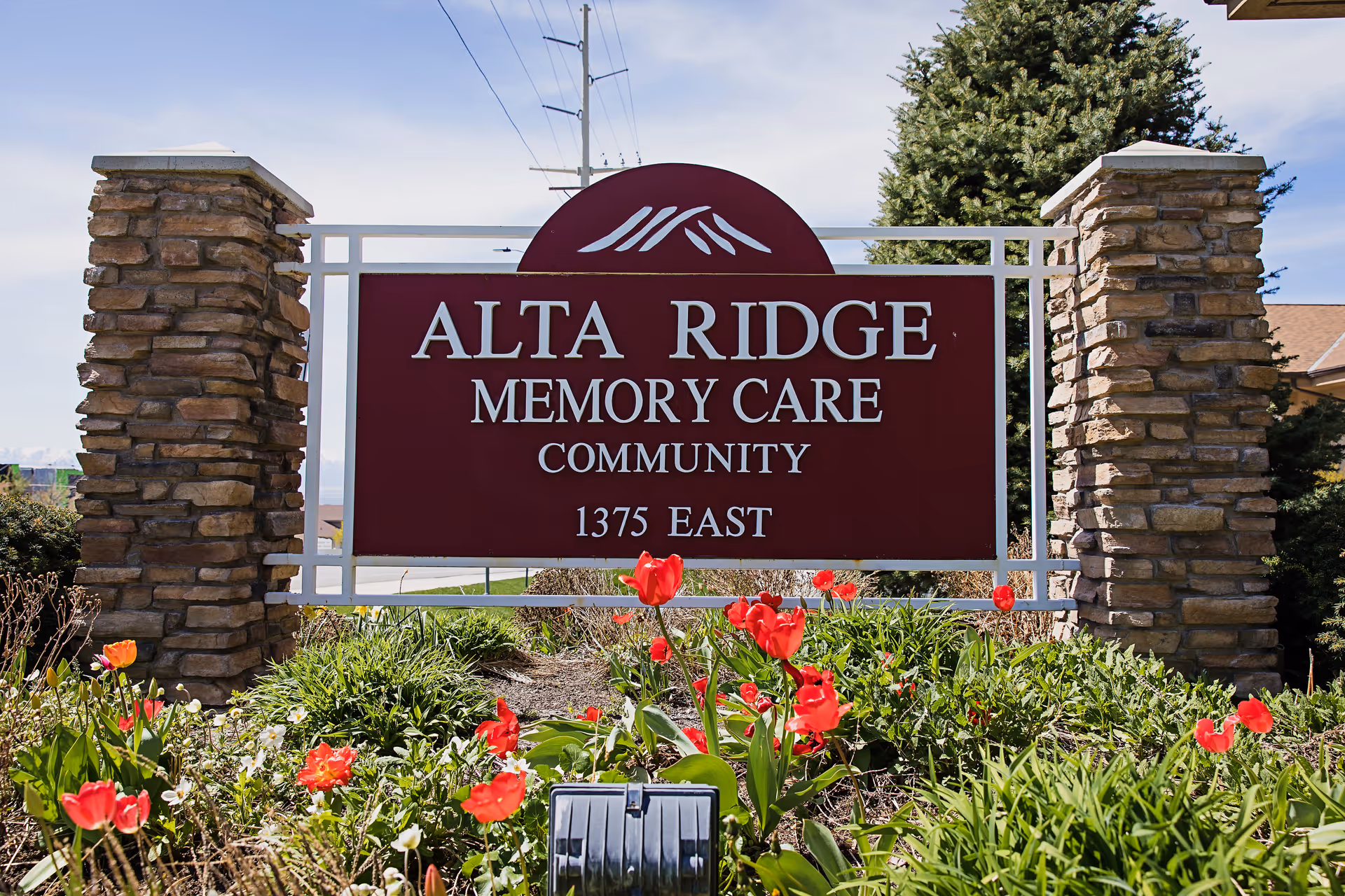 A large maroon sign with white text that reads 'ALTA RIDGE MEMORY CARE COMMUNITY 1375 EAST' mounted between two stone pillars, surrounded by green plants and red flowers under a partly cloudy sky.