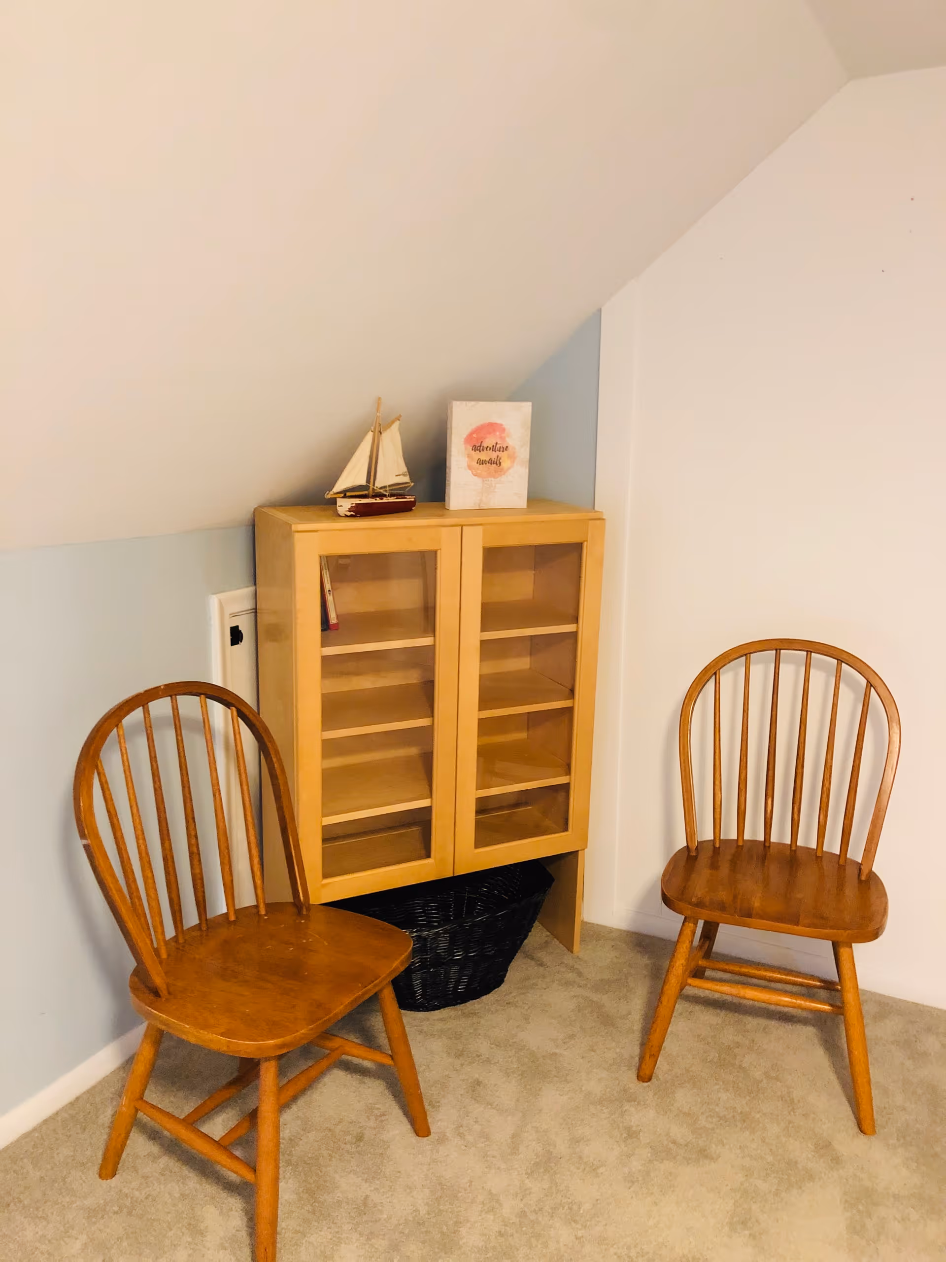 Two wooden chairs placed on a carpeted floor next to a small wooden cabinet with glass doors. On top of the cabinet, there is a small decorative sailboat and a framed picture with the words 'adventure awaits'. The walls are painted light blue and white, with a slanted ceiling above.