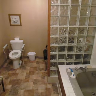 A bathroom with a toilet, a small trash bin, a framed picture on the wall, and a bathtub with glass block partition. The floor has a patterned tile design.