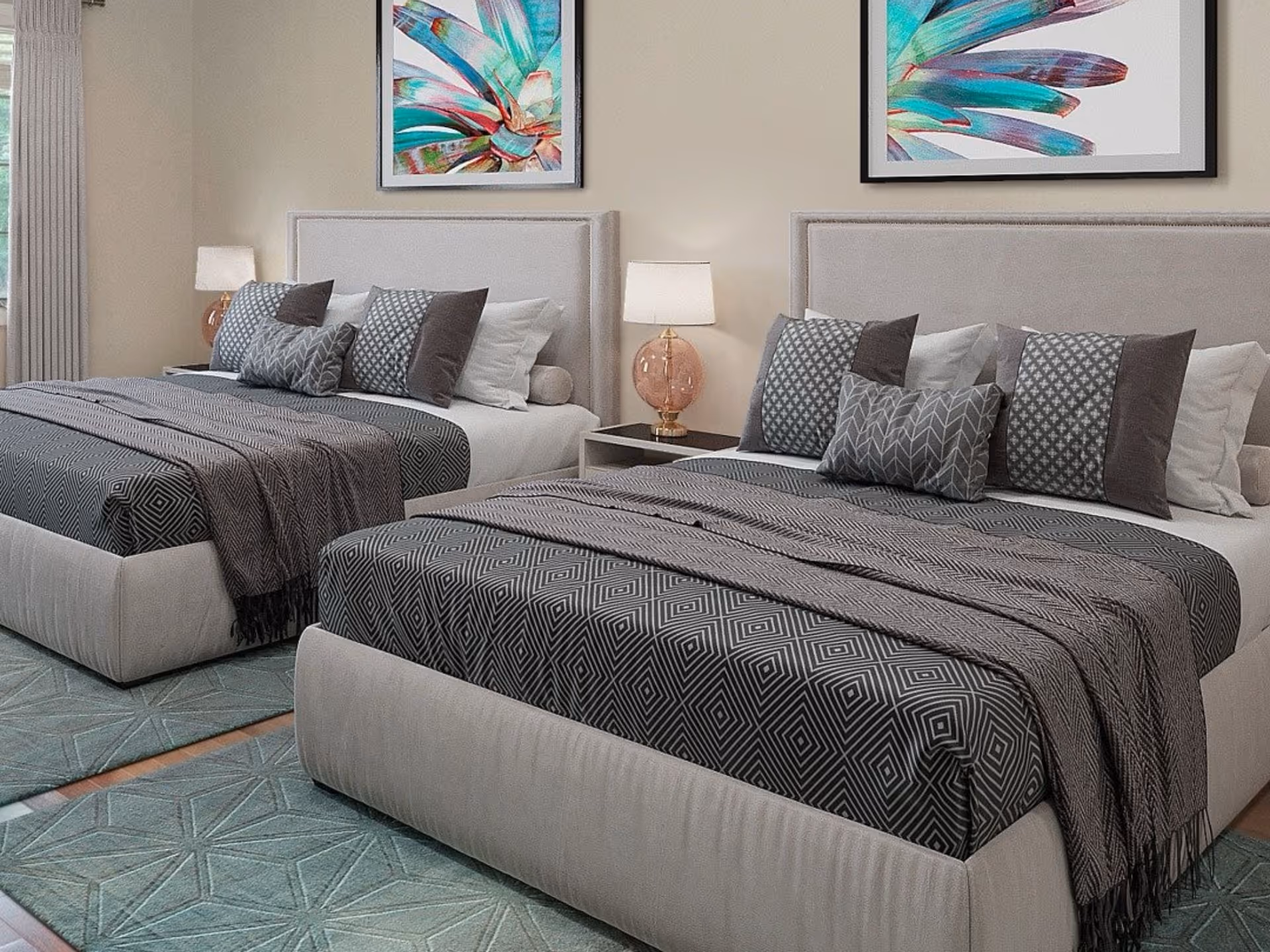 A bedroom with two neatly made beds featuring gray patterned bedding and multiple pillows. Each bed has a beige upholstered headboard. Between the beds is a nightstand with a lamp that has a pink glass base. Above each bed hangs a colorful abstract painting with shades of teal, pink, and purple. A window with beige curtains is visible on the left side of the room.