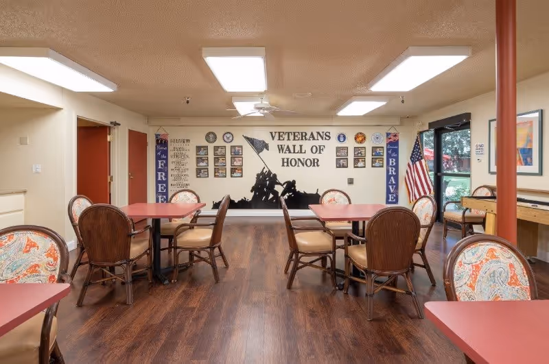 Dining/activity room with tables and chairs facing a 'Veterans Wall of Honor' mural and an American flag.