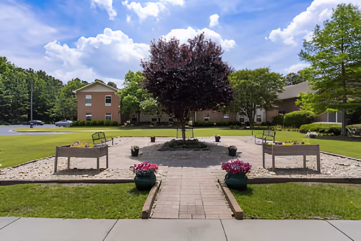 Outdoor garden area with a paved walkway leading to a central tree surrounded by benches and flower pots. The garden is bordered by green grass and trees, with buildings visible in the background under a partly cloudy sky.