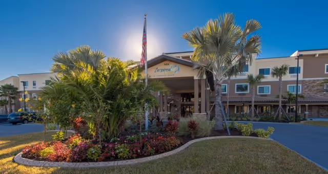 Front entrance of Inspired Living Lakewood Ranch with a landscaped circular planter, palm trees, and an American flag under a sunny blue sky.