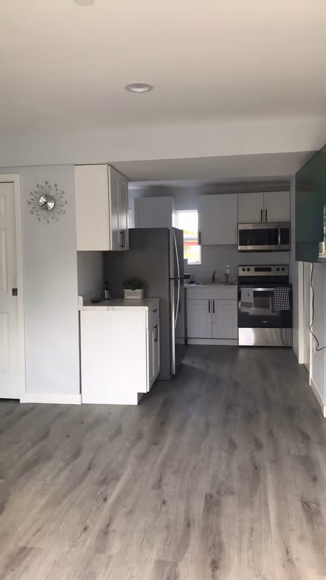 A modern kitchen with white cabinets, a stainless steel refrigerator, oven, and microwave. There is a small plant on the counter and a clock on the wall. The floor is a light wood laminate.