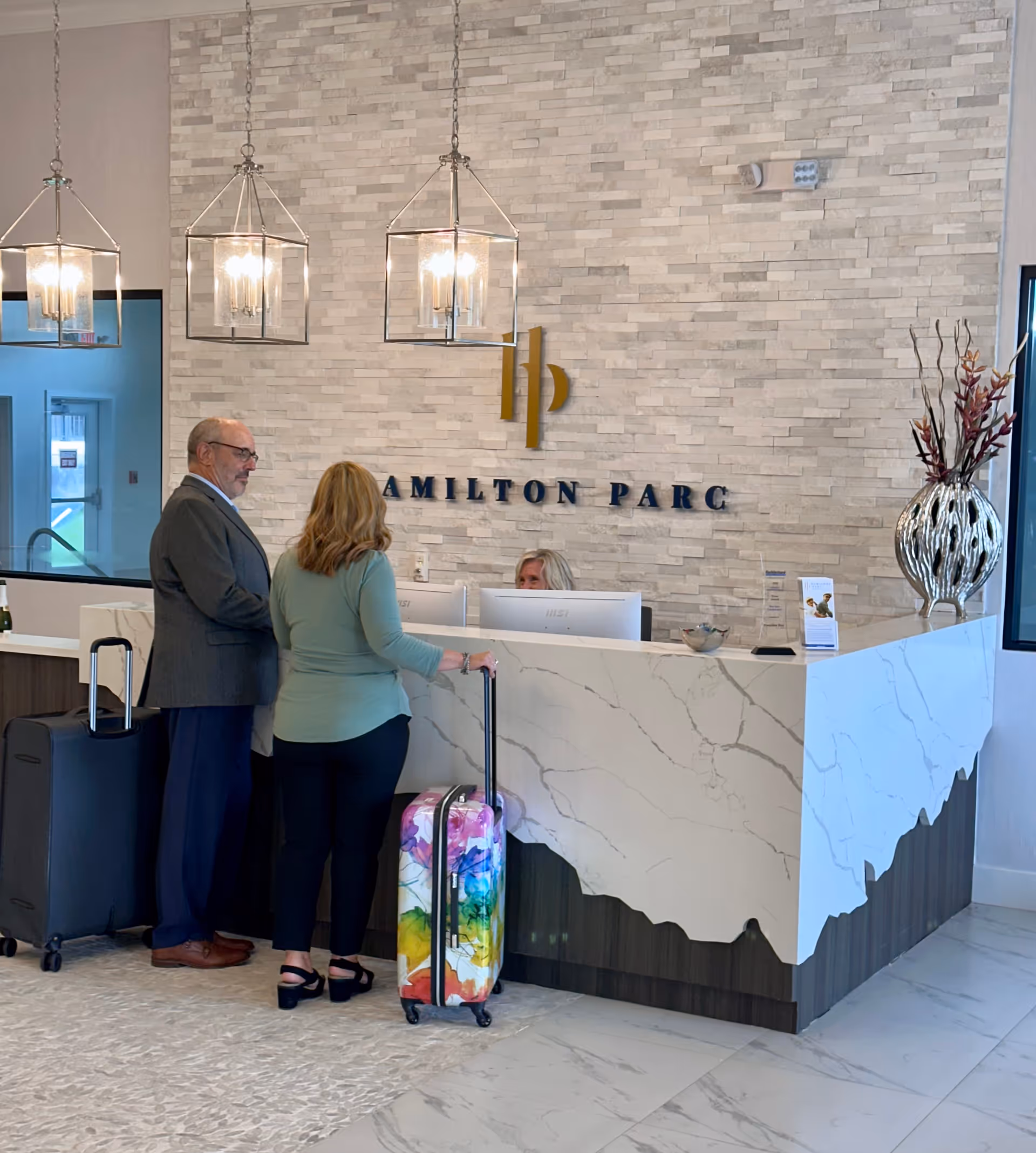 Two people with suitcases standing at a modern reception desk with a receptionist behind the counter. The wall behind the desk has a stone tile design with the text 'Hamilton Parc' and a gold logo. Three pendant lights hang from the ceiling above the desk, and a decorative vase with tall branches is on the right side of the counter.