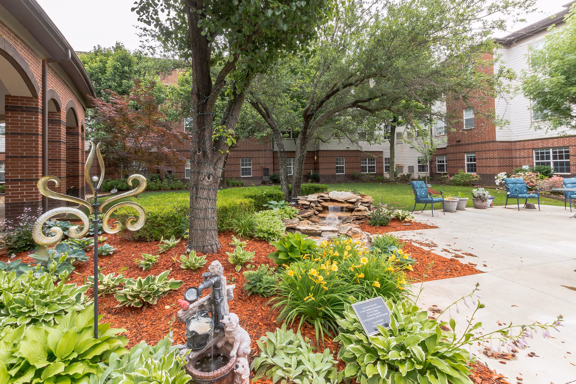 Landscaped courtyard with flower beds, a small rock waterfall, patio seating and a brick senior living building in the background.