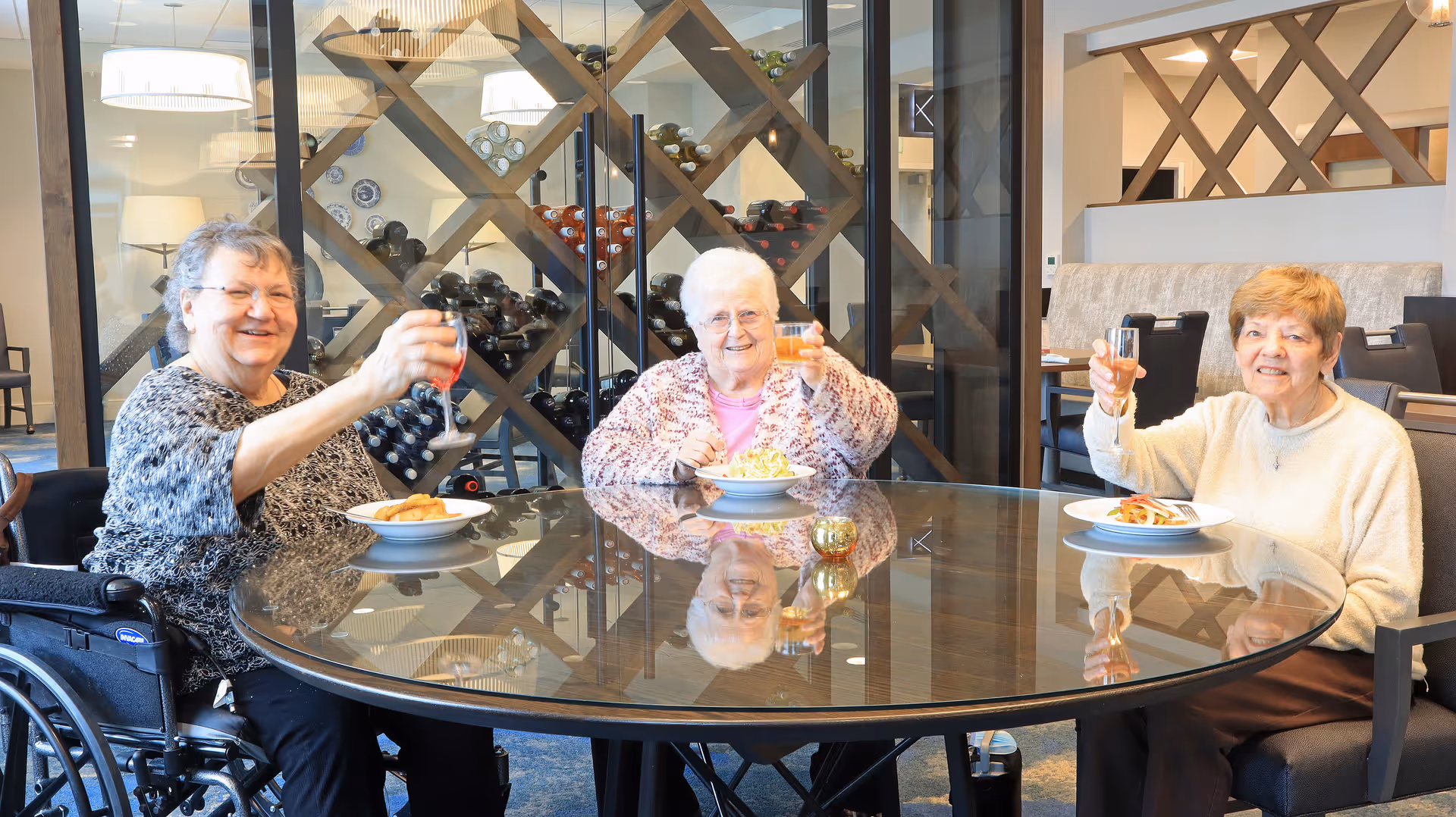 Three elderly women sitting around a round glass table in a dining area, each holding a glass and smiling. Behind them is a wine rack filled with bottles. The setting appears warm and inviting with modern decor.