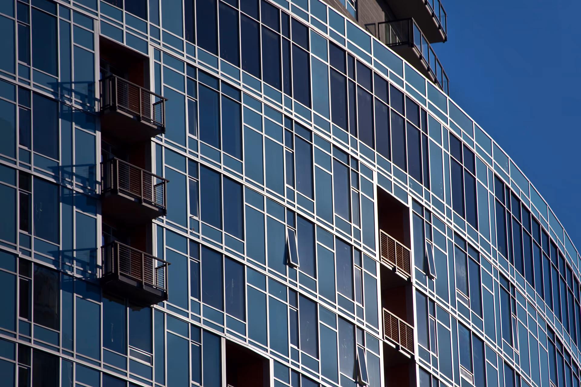 Curved glass high-rise building facade with multiple small balconies and reflective windows against a blue sky.