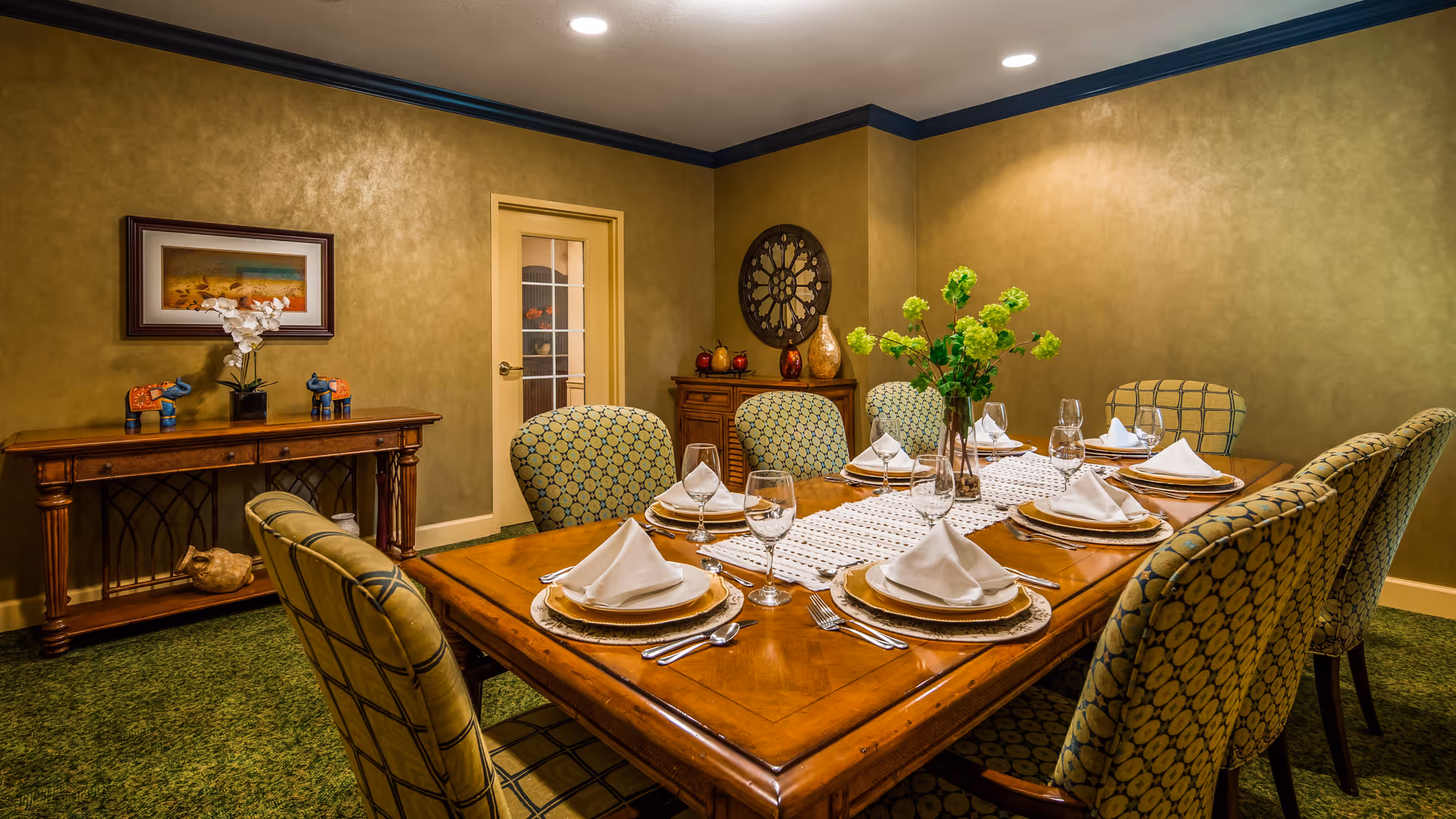 A formal dining room with a wooden table set for eight people. Each place setting includes a plate with a folded white napkin, a wine glass, and silverware. The chairs around the table have patterned upholstery. The room has green carpet, beige walls, and decorative items including a vase with green flowers on the table, framed artwork on the wall, and a wooden sideboard with decorative objects.