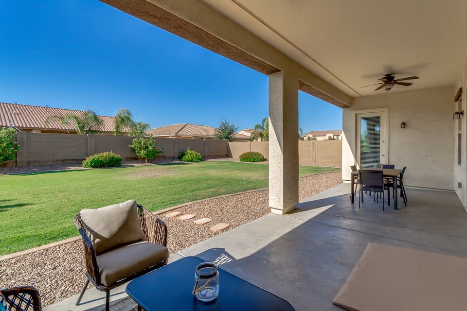 Covered patio area with outdoor seating including a cushioned chair and a table with a glass lantern, overlooking a spacious backyard with green grass, small bushes, and a tall privacy fence under a clear blue sky.