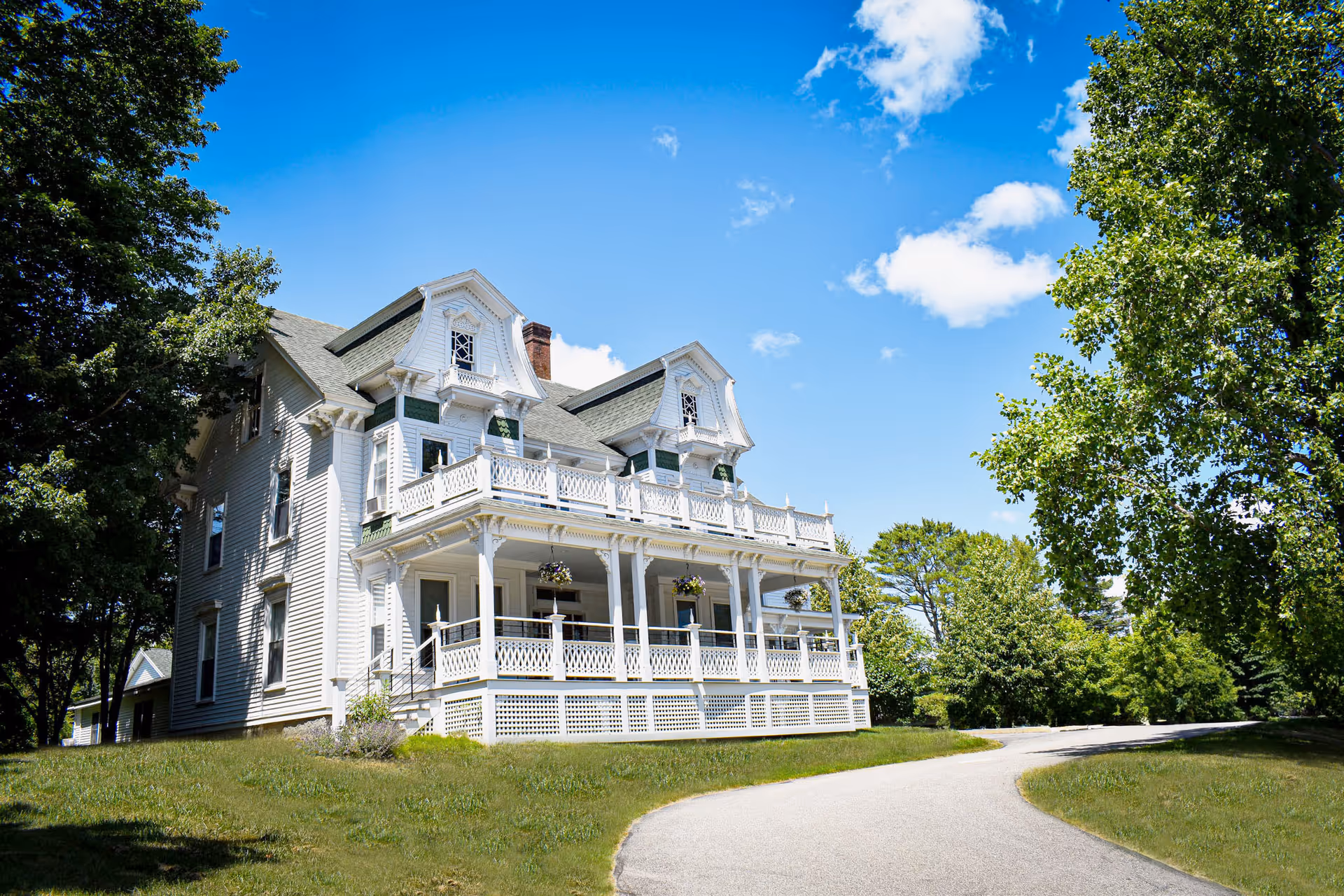 Large white Victorian-style manor with a wraparound porch on a grassy lawn under a bright blue sky.