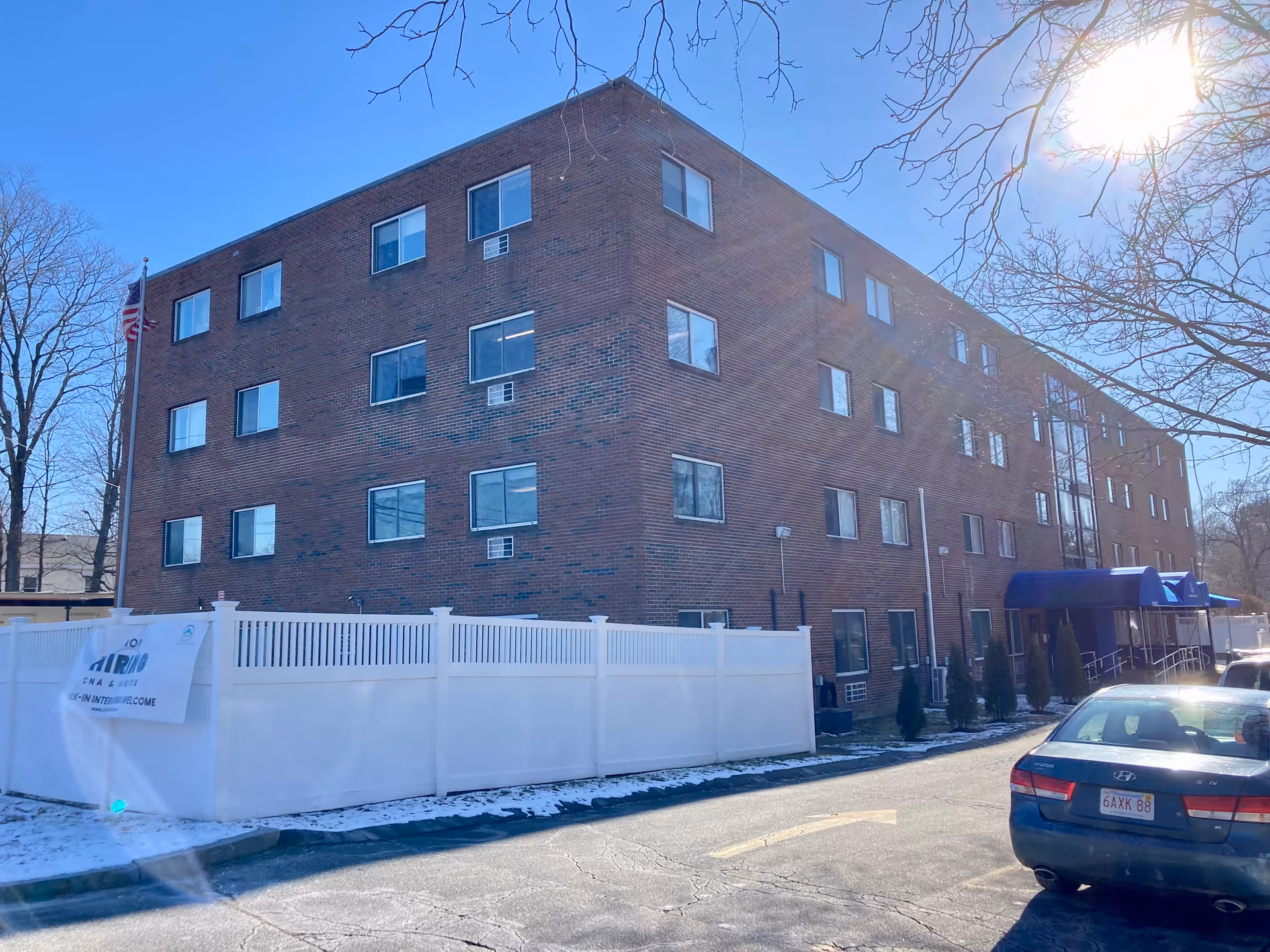 Exterior view of a four-story brick building with multiple windows, a white fence, a flagpole with an American flag, and a blue awning over the entrance. There is a car parked on the right side of the image and some snow on the ground. The sky is clear and sunny.