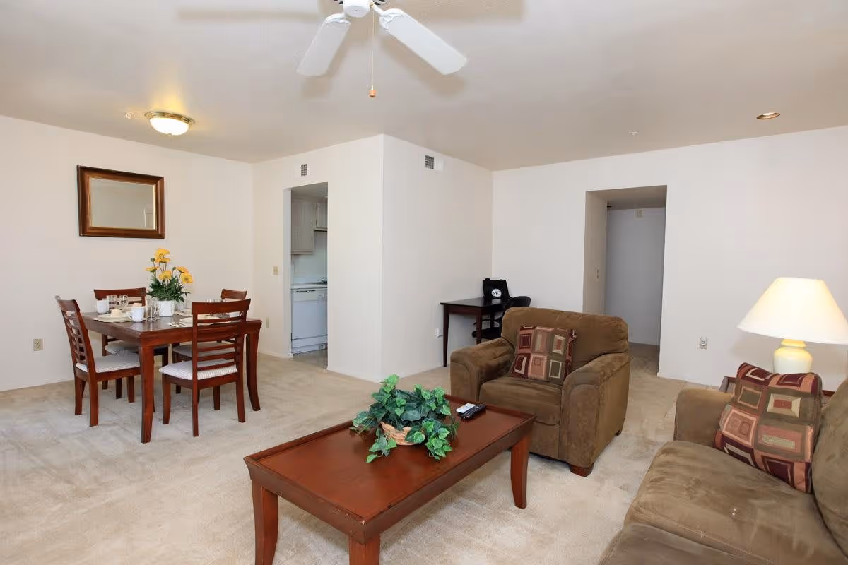 A living room and dining area in a senior living facility with beige carpet and white walls. The living room has a brown sofa and armchair with patterned cushions, a wooden coffee table with a green plant centerpiece, and a table lamp. The dining area has a wooden table set with four chairs, a flower arrangement, and place settings. A ceiling fan and light fixture are visible on the ceiling. A small desk with a chair is positioned against the wall near an open doorway leading to a kitchen.