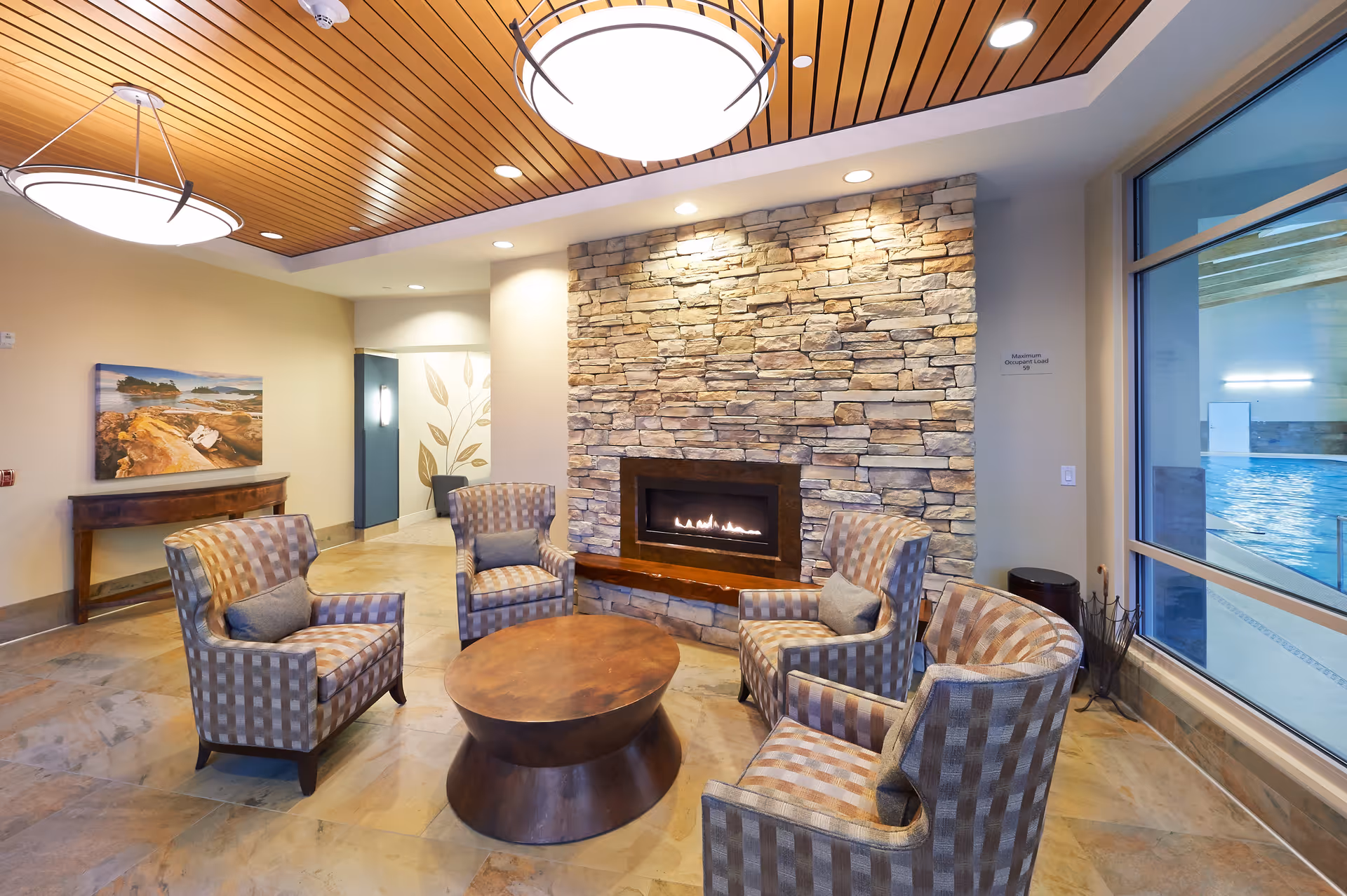 A cozy seating area with five patterned armchairs arranged around a round wooden coffee table in front of a stone fireplace with a fire burning. The room has a wooden ceiling with modern circular light fixtures, a large window showing an indoor pool area, and a wall with a landscape painting and a decorative panel with leaf designs.