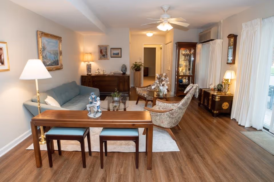 A cozy living room with wooden flooring, featuring a light blue sofa, two patterned armchairs, a wooden coffee table, and a wooden console table with two stools underneath. The room is decorated with framed paintings on the walls, a floor lamp, table lamps, a ceiling fan, and a glass display cabinet. Large windows with white curtains allow natural light to enter the space.