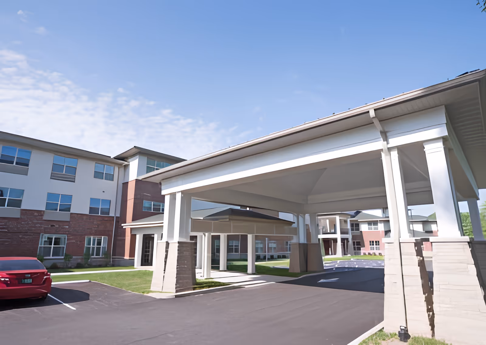 Exterior view of a senior living facility with a covered entrance supported by white columns. The building has multiple windows and a combination of brick and light-colored siding. A red car is parked in the parking lot near the entrance under a clear blue sky.