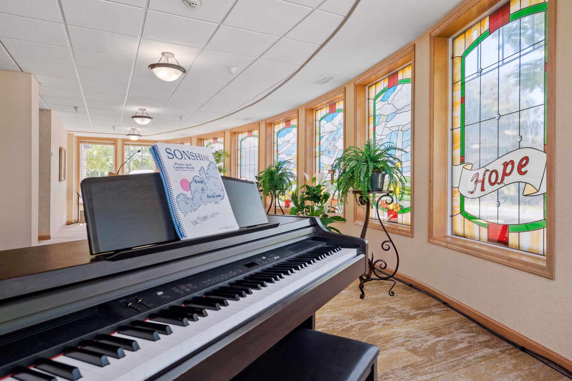 A digital piano in a bright curved interior corridor with stained-glass windows (one reading 'Hope') and potted plants.