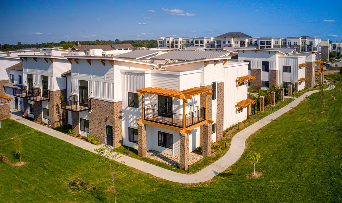Aerial view of a modern senior living facility with multiple two-story buildings featuring balconies with wooden pergolas, surrounded by well-maintained green lawns and a paved walkway under a clear blue sky.